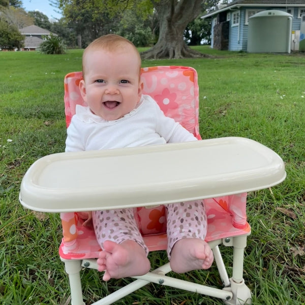 Bloom Portable Baby Highchair with detachable tray. Comes in a carry bag. For kids aged 6M-3Y. Bloom Pattern is pink with pink and neutral toned flowers. Image depicts a happy child sitting in the chair in a park.