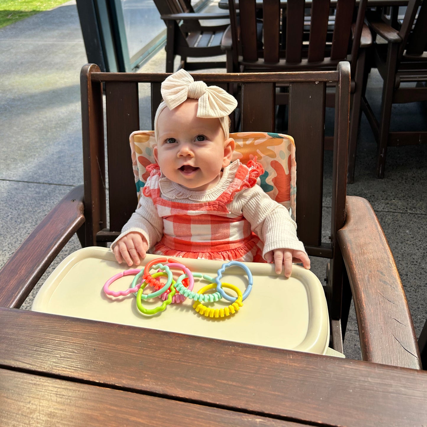 Ziggy Portable Baby Highchair with detachable tray. Comes in a carry bag. For kids aged 6M-3Y. Ziggy Pattern has neutral background with dark blue, orange, peach, yellow and light blue blobs. Image depicts a happy baby sitting in the chairs converting a cafe chair into a highchair.