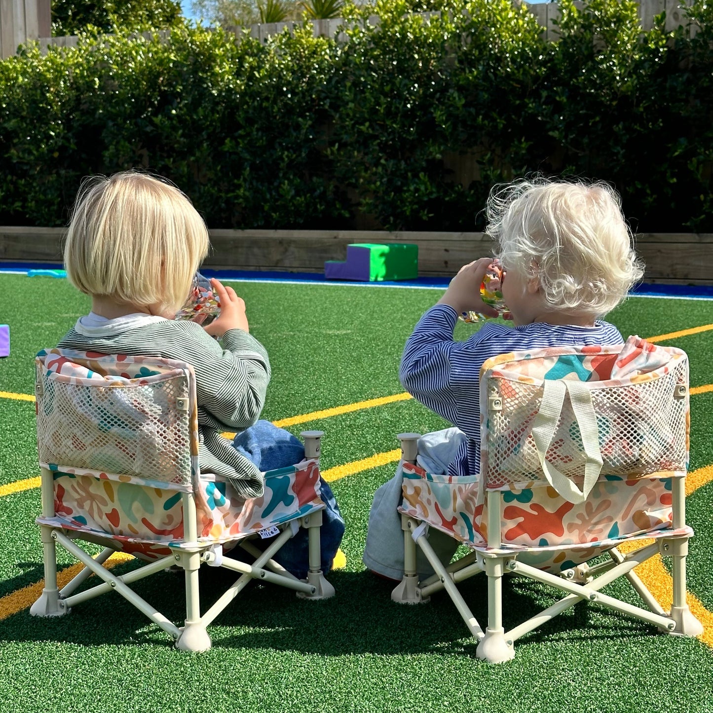 Ziggy Portable Baby Highchair with detachable tray. Comes in a carry bag. For kids aged 6M-3Y. Ziggy Pattern has neutral background with dark blue, orange, peach, yellow and light blue blobs. Image depicts two twin boys sitting in the chairs at a playground, from behind.