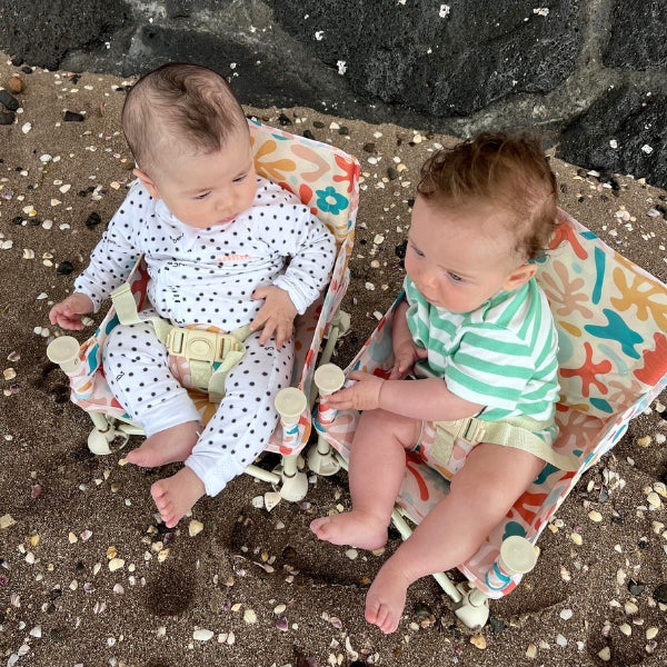 Ziggy Portable Baby Highchair with detachable tray. Comes in a carry bag. For kids aged 6M-3Y. Ziggy Pattern has neutral background with dark blue, orange, peach, yellow and light blue blobs. Image depicts two happy babies sitting in the chairs at the beach from above.