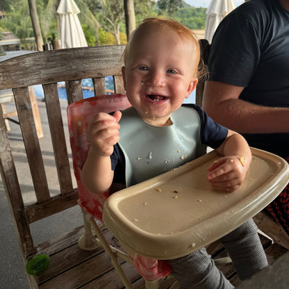 Bloom Portable Baby Highchair with detachable tray. Comes in a carry bag. For kids aged 6M-3Y. Image depicts a happy baby sitting in the chairs converting a cafe chair into a highchair.