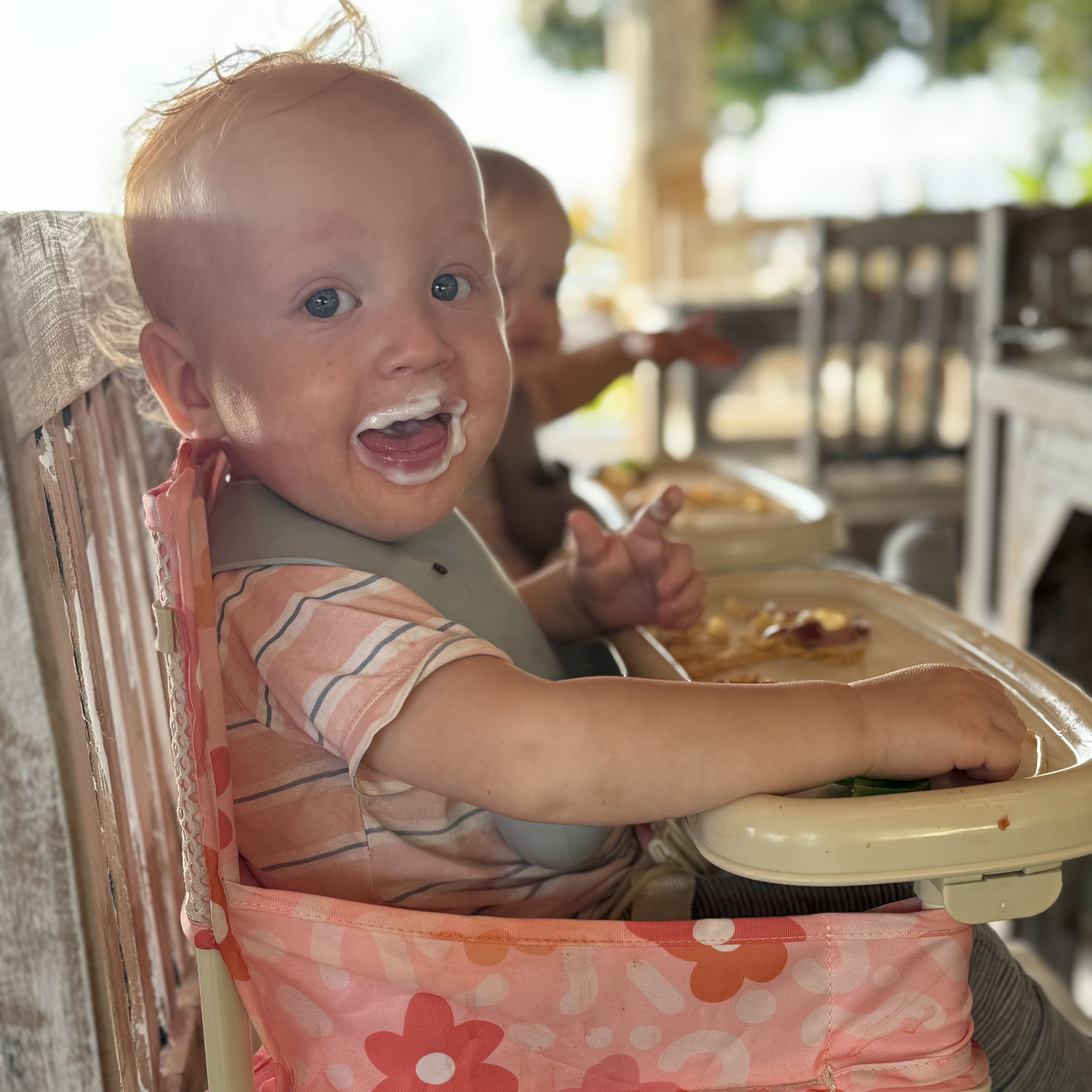 Bloom Portable Baby Highchair with detachable tray. Comes in a carry bag. For kids aged 6M-3Y. Image depicts a happy baby sitting in the chairs converting a cafe chair into a highchair.